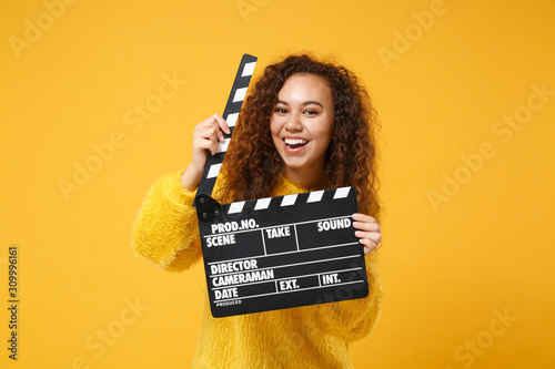 Cheerful young african american girl in fur sweater posing isolated on yellow orange background in studio. People lifestyle concept. Mock up copy space. Holding classic black film making clapperboard.
