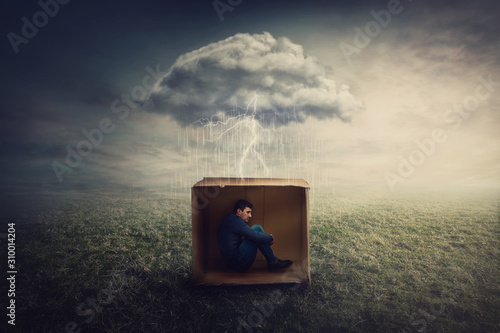 Surreal concept with a scared guy shelters inside a cardboard box. Introvert man caged by own fears as a thunderstorm cloud trapped him under the rain. Mysterious storm as emotional crisis symbol.