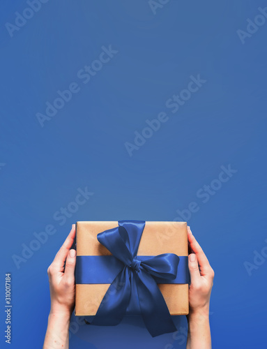 Female hands hold gift box on deep blue background with copy space for design. Caucasian girl hands holding gift box in craft wrapping paper with fashion color 2020 blue satin ribbon. Vertical.