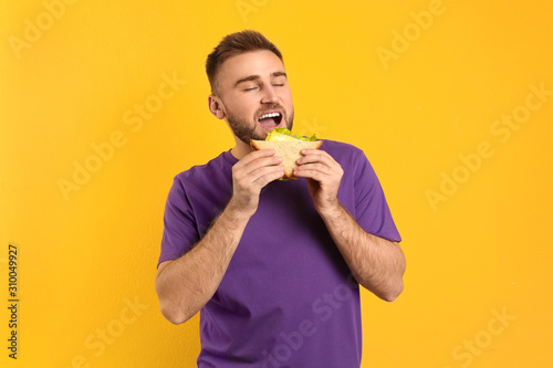 Young man eating tasty sandwich on yellow background