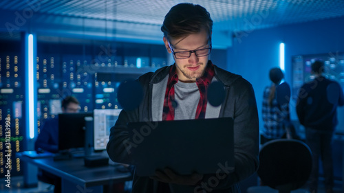 Portrait of a Smart Focused Young Man Wearing Glasses Holds Laptop. In the Background Technical Department Office with Specialists Working and Functional Data Server Racks