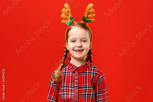 Funny happy baby in a red dress and deer horns on his head on a colored background. The little girl is laughing