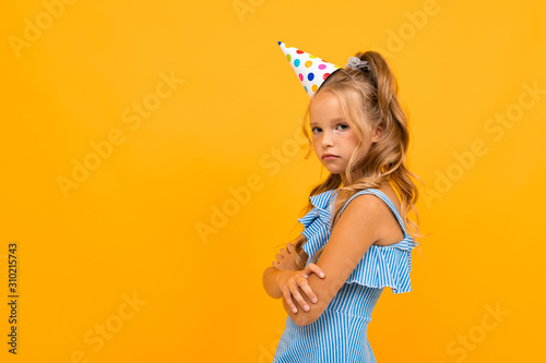offended girl in a festive cap stands sideways with folded arms on a yellow background