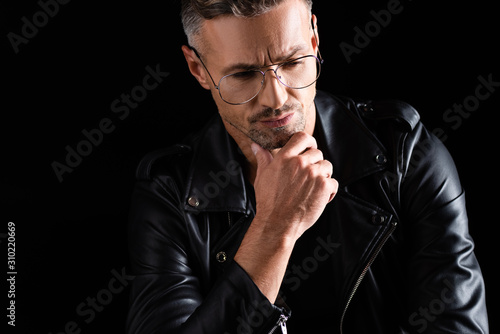 Pensive man in sunglasses and biker jacket with hand by chin isolated on black