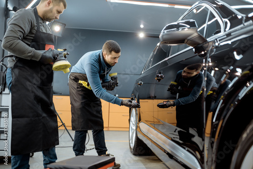 Car service workers examining glossy vehicle body coating for scratches after the polishing procedure at the service station. Car detailing concept