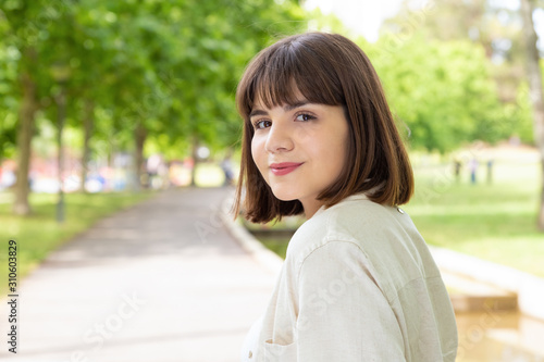 Attractive young woman smiling at camera outdoors. Portrait of beautiful cheerful young woman standing and looking at camera in park. Facial expression concept