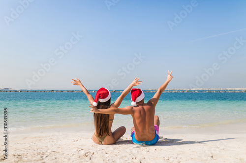 Young couple in love in santa hat have fun with raised hands on the beach. Rear view.