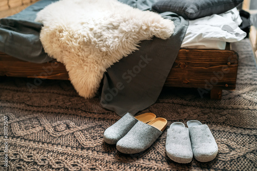 The two pair of gray home slippers near the wooden bed on the "knitted floor" in the cozy bedroom. Home sweet home concept image.