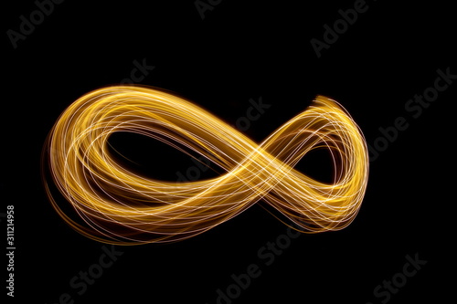 Long exposure photograph of an infinity loop in gold neon colour in an abstract swirl, parallel lines pattern against a black background. Light painting photography.