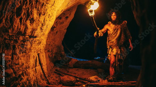 Primeval Caveman Wearing Animal Skin Exploring Cave At Night, Holding Torch with Fire Looking at Drawings on the Walls at Night. Neanderthal Searching Safe Place to Spend the Night