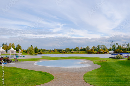 Landscape. View of the beautiful lake and small ornamental ponds covered with paving tiles, lawn with grass, ornamental shrubs and trees, tents and loungers for recreation in the park.
