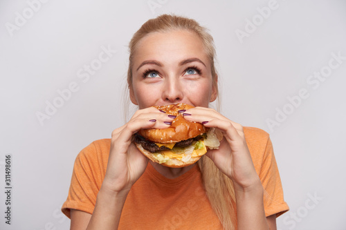 Portrait of pleased young lovely blonde woman with casual hairstyle eating fresh hamburger with great appetite and looking cheerfully upwards, posing over white background