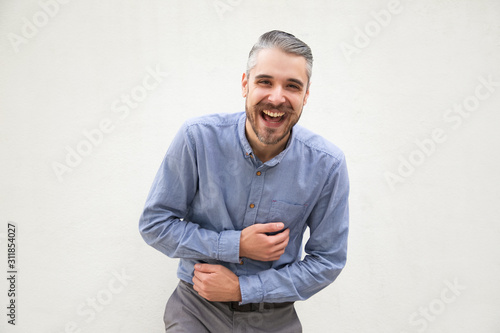 Happy excited man laughing out loud, touching stomach. Grey haired young man in blue casual shirt posing isolated over white background. Fun or laughter concept