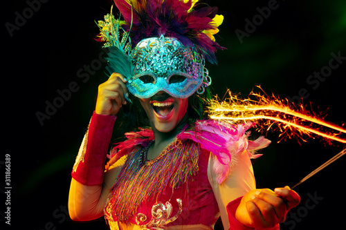 Beautiful young woman in carnival mask and stylish masquerade costume with feathers and sparklers in colorful lights on black background. Christmas, New Year, celebration. Festive time, dance, party.