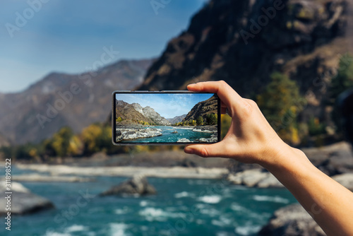 Female hand with smartphone close-up. Taking photo on smartphone while traveling. Delightful mountain landscape, stormy river, rocks and blue sky.