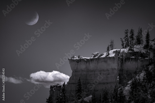 Black and white, fine art scene of a half moon setting at night over a rock cliff in Bryce Canyon National Park, Utah. 