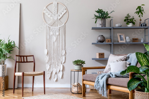 Stylish interior of living room at cozy apartment with brown sofa, coffe table, bookstand, a lot of plants, macrame and elegant accessories. Beige and japandi concept. Modern home staging. Template.