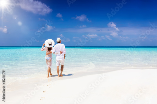 A hugging honeymoon couple walks down a tropical beach with turquoise sea and sunshine 