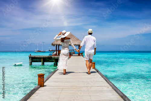 A happy couple in white summer clothing on vacation walks along a wooden pier over tropical, turquoise ocean in the Maldives, Indian Ocean