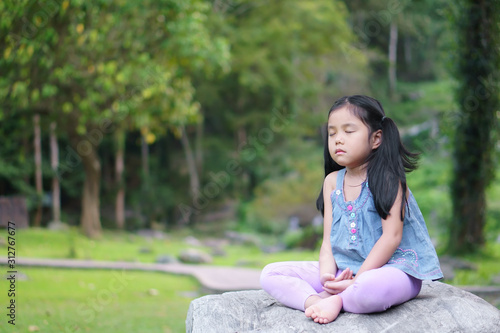 Asian child cute or kid girl close eye mindfulness meditating with breath and sit on stone or rock for peace and relax or yoga on nature green garden and tree at temple park or church outdoor