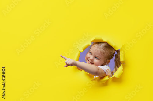 Little surprised child looking, peeping through the bright yellow paper hole. Showing hand to side. Advertise childrens goods. Copy space for text.