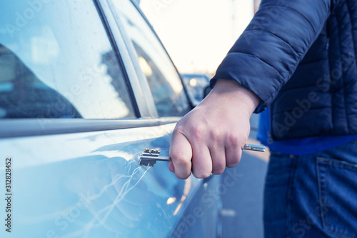 Bad guy scratching the car door with a key in the parking lot on the street. Damage of property from revenge for treason or betrayal, or threat. Auto insurance fraud or vandalism.