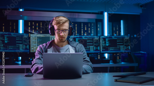Portrait of Software Developer / Hacker / Gamer Wearing Glasses and Headset Sitting at His Desk and Working / Playing on Laptop. In Background Dark Neon High Tech Environment with Multiple Displays.