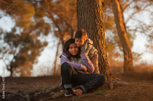 A big sister and young brother sitting near a tree on a hill and hugging each other while enjoying a sunset.