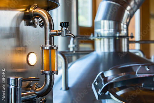 sight glass full of golden beer on stainless steel pipe with hose bib faucet valve and tri clamps selective focus, brewery tank and vessel background