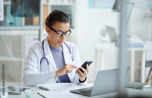 Serious female doctor in white coat sitting at her workplace and typing a message on her mobile phone at office