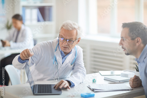 Senior doctor in eyeglasses sitting at the table pointing at laptop computer and discussing illness together with his patient