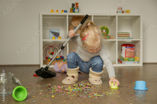 Adorable blonde toddler girl playing with broom, cleaning colorful confetti from the floor