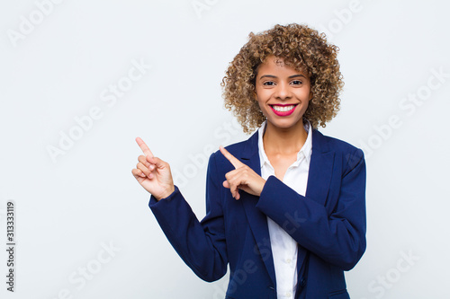 young woman african american smiling happily and pointing to side and upwards with both hands showing object in copy space against flat wall