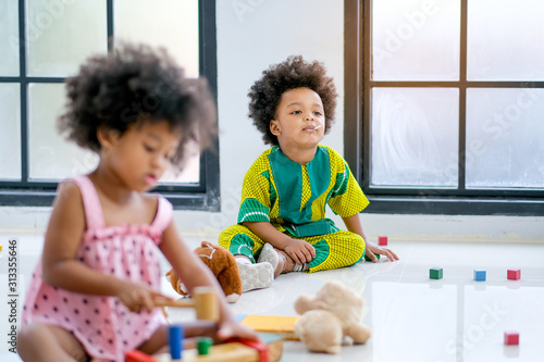 Young African little boy is inattentive during play toys and sit on back of the other little girl in living room with day light.