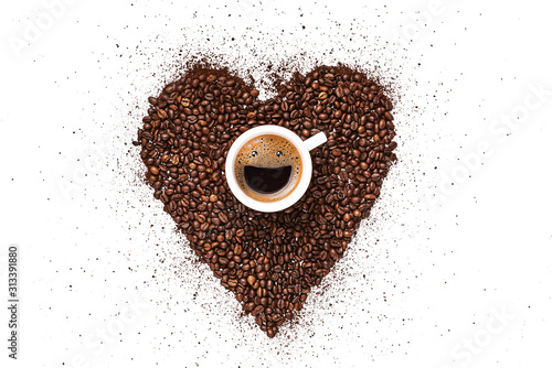 heart made from coffee beans and ground coffee on a white background. in the center is a cup, in a cup from coffee foam a happy smiling face. invigorating drink concept