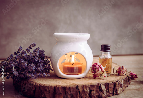 Vintage style picture of white ceramic candle aroma oil lamp with essential oil bottle and dry flower petals on natural pine wood disc, dry background with copy space.