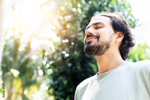 A bearded man is meditating outdoor in the park with face raised up to sky and eyes closed on sunny summer day. Concept of meditation, dreaming, wellbeing healthy lifestyle