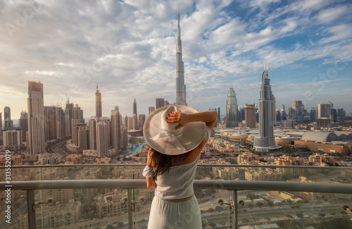 Woman with a white hat is standing on a balcony in front of the skyline from Dubai Downtown