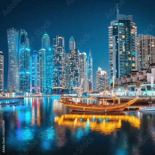 Multicolored lights of the night city in the Dubai Marina district. Stylized ancient Arabic ship Abra Dhow with lights in the foreground.