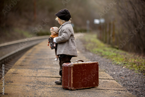 Adorable boy on a railway station, waiting for the train with suitcase and beautiful vintage doll...