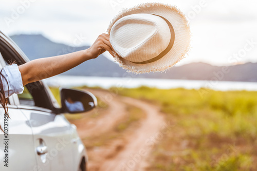 Happy woman hand holding hat outside open window car with meadow and mountain lake background. People lifestyle relaxing as traveler on road trip in holiday vacation. Transportation and travel concept