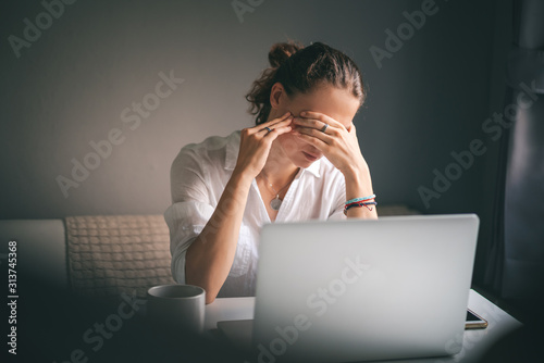 Young pretty woman in a white shirt sits at a table in front of a laptop, her eyes and head ache, workaholism, stress burnout