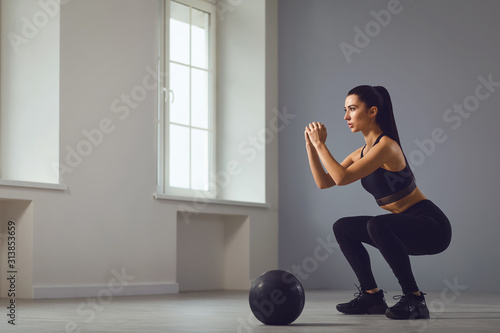 Squat exercises. Girl in black sportswear with dumbbells in her hands doing squats in a room.