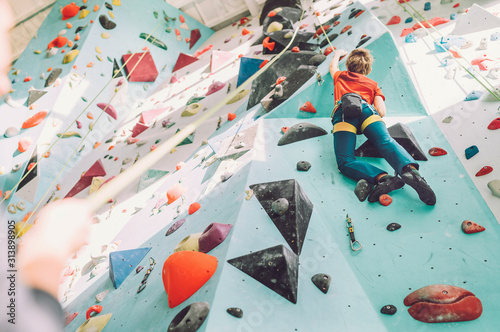 Teenager boy at indoor climbing wall hall. Boy is climbing using a top rope and climbing harness and somebody belaying him from floor. Active teenager time spending concept image.
