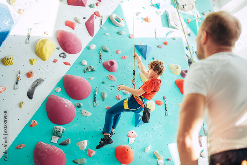 Father and teenager son at indoor climbing wall hall. Boy is hanging on the rope using a climbing harness and daddy belaying him on the floor using a belay device. Happy parenting concept image.