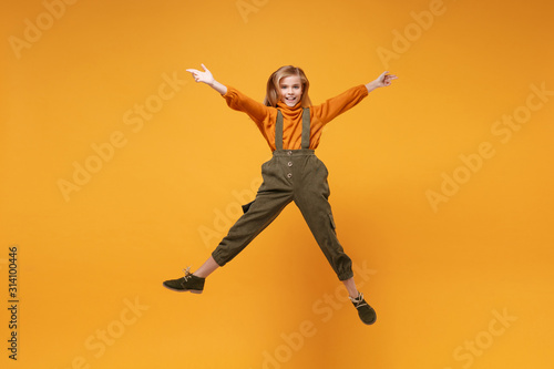 Smiling little kid girl 12-13 years old in turtleneck, jumpsuit isolated on orange yellow wall background in studio. Childhood lifestyle concept. Mock up copy space. Jumping, spreading hands and legs.