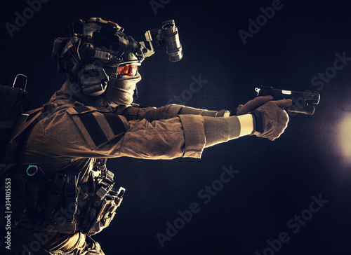 Modern soldier, counter terrorist squad fighter in combat uniform, helmet and tactical radio headset, aiming service pistol in darkness, shooting with handgun, studio portrait on dark background