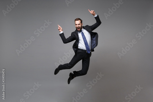 Happy young business man in classic black suit shirt tie posing isolated on grey background. Achievement career wealth business concept. Mock up copy space. Jumping, point index fingers up, screaming.