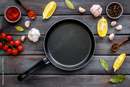 Cooking mockup. Frying pan among spices and vegetables on dark wooden desk top-down copy space
