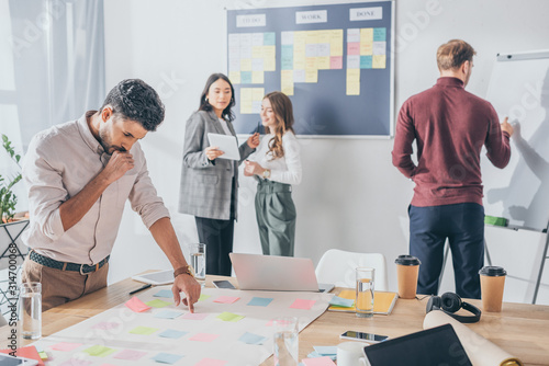 selective focus of pensive mixed race scrum master pointing with finger at sticky note near coworkers